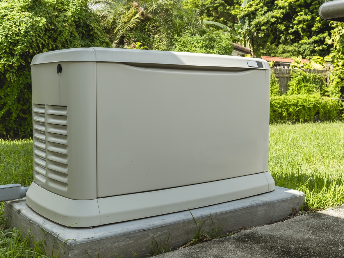 Home standby generator on a concrete pad in a lush backyard, surrounded by greenery and lawn.